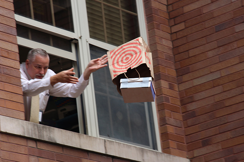 A man wearing a white shirt and gold tie tosses a box out a second floor window. The box has a paper bag attached to the top like a parachute.