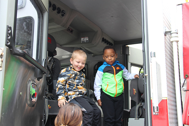 Two elementary boys sit on seats inside of a fire truck, smiling.