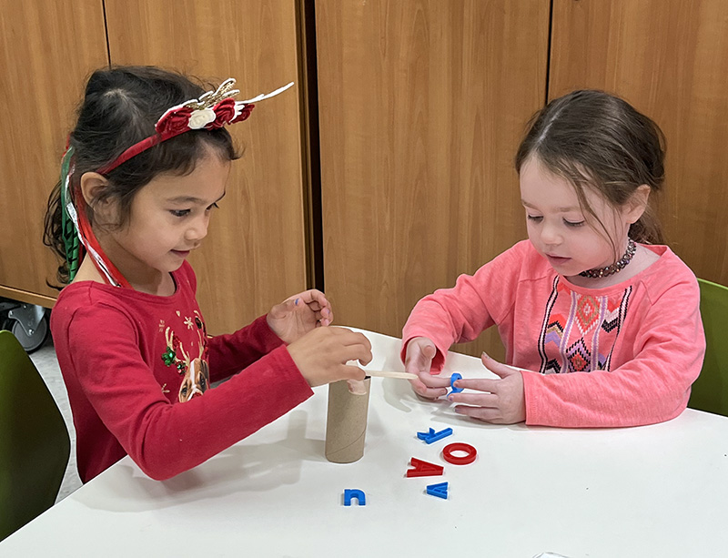 Two kindergarten girls sit at a table and build a tree with branches. There are plastic letters on the table.