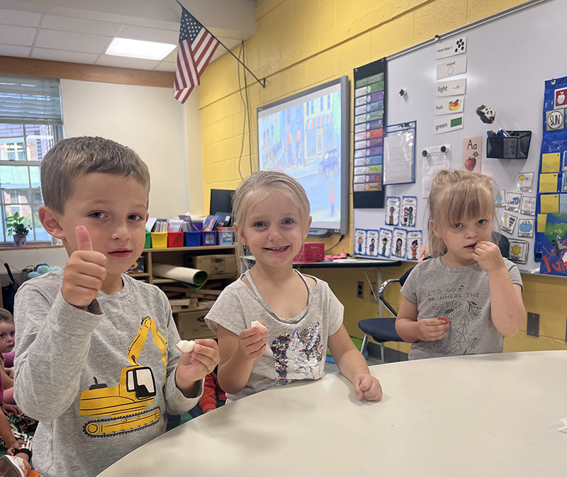 Three pre-K students eat pieces of apple. The boy on the left has his thumb up and holds a piece of apple in his other hand. The girl in center is holding up her piece of apple and smiling. The girl on the right is eating her piece of apple.