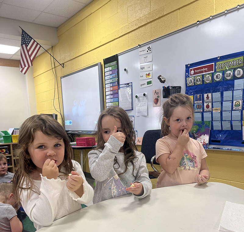 Three pre-K girls sit at a table. Two are eating a piece of apple and the girl in the center is giving a thumbs up.