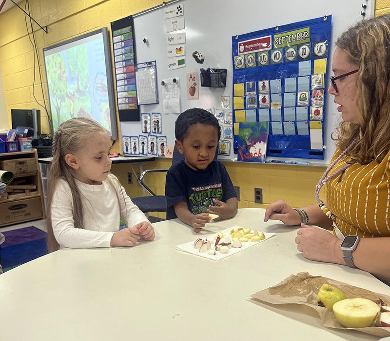 Two pre-K students choose pieces of apple from a plate of them on a table. There is a woman there pointing to the apples.