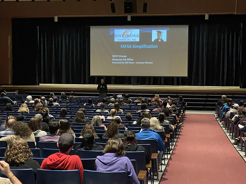 Students and adults sit in an auditorium with a large screen in front and a man speaking.