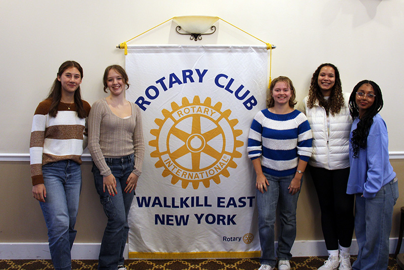 Five high school young woman - two on the left and three on the right - flank a Rotary Club banner.