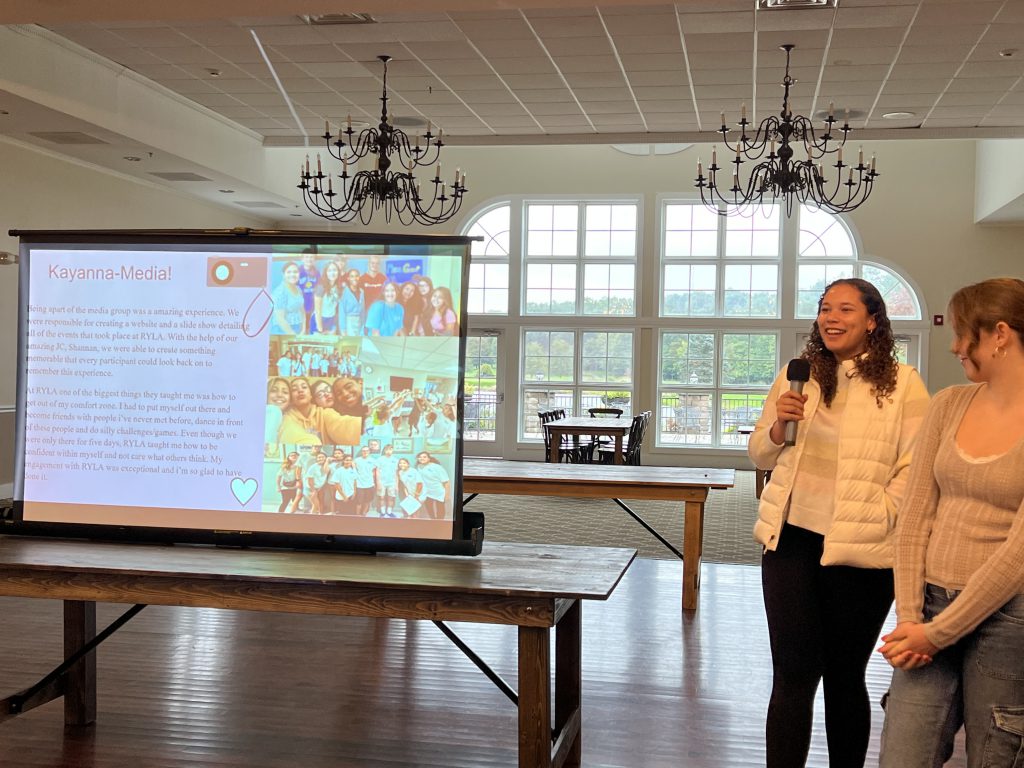 A high school young woman holds a microphone and presents to a group of people from what is displayed on a screen.