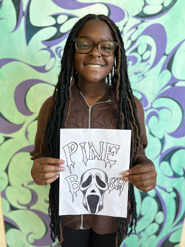 A high school girl smiles broadly. She has long dark hair and is wearing glasses. She is holding a drawing that says Pine Boosh with a skull's eyes serving as the two o's in boosh.