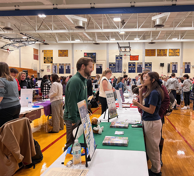 A man on one side of a table speaks with a young woman on the other side. They are surrounded by many other people.