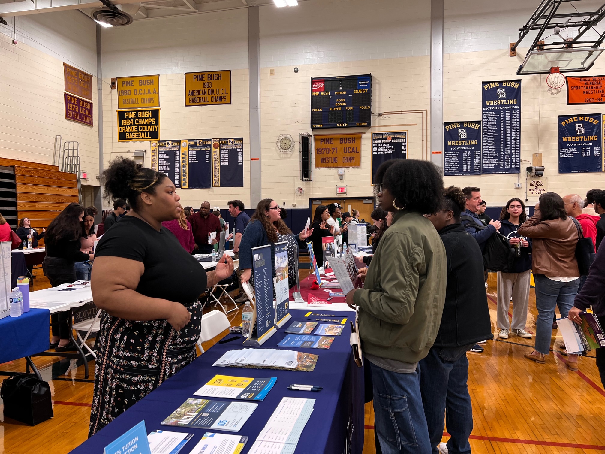 A woman on one side of a table talks with a young woman on the other side. The table has a blue table cloth and many pamphlets on it. There are other people in the background.