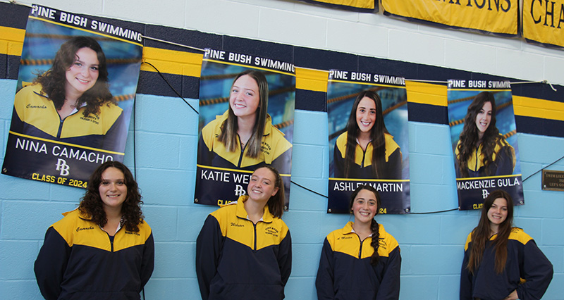 Four senior young women smile and stand in front of their banners that are hanging on the wall. The banners have their photos on them.