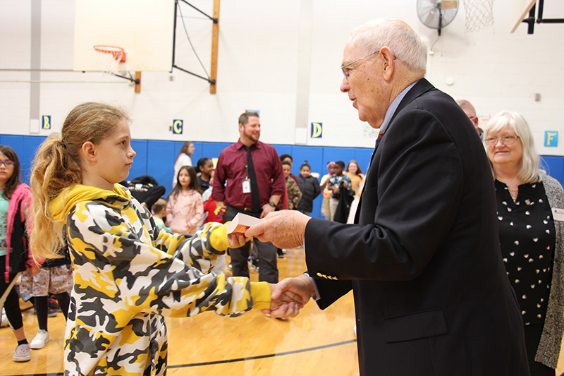 A man in a black suit jacket shakes hands and gives a red dictionary to a young girl in a multi-colored sweatshirt.