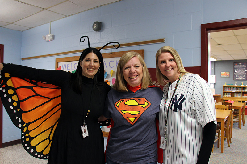 Three women are dressed in costume - a buttefrly on the left, supergirl in the center, and a NY Yankeed baseball player on the right. All are smiling.