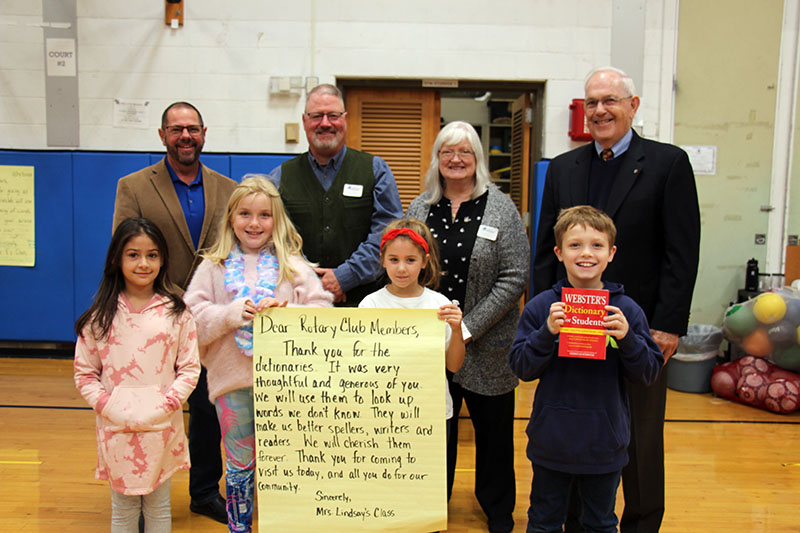 A group of four third-grade kids stand in front while four adults stand in back. The boy on the right holds a red dictionary. The two girls in the center hold a large thank you note and the girl on the left smiles.