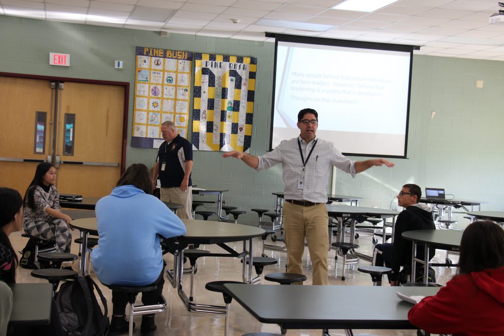 A man stands in the center of a room with his arms out wide talking. There is a man in the background and students in seats arll around.