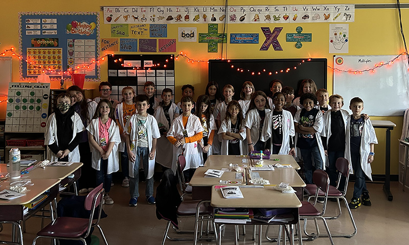 A large group of fourth-graders all wearing white lab coats, stand in front of a classroom and smile.