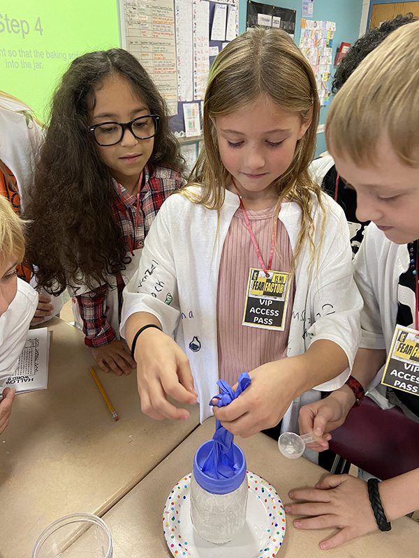 Three fourth-grade students work with a blue rubber glove over a clear jar.