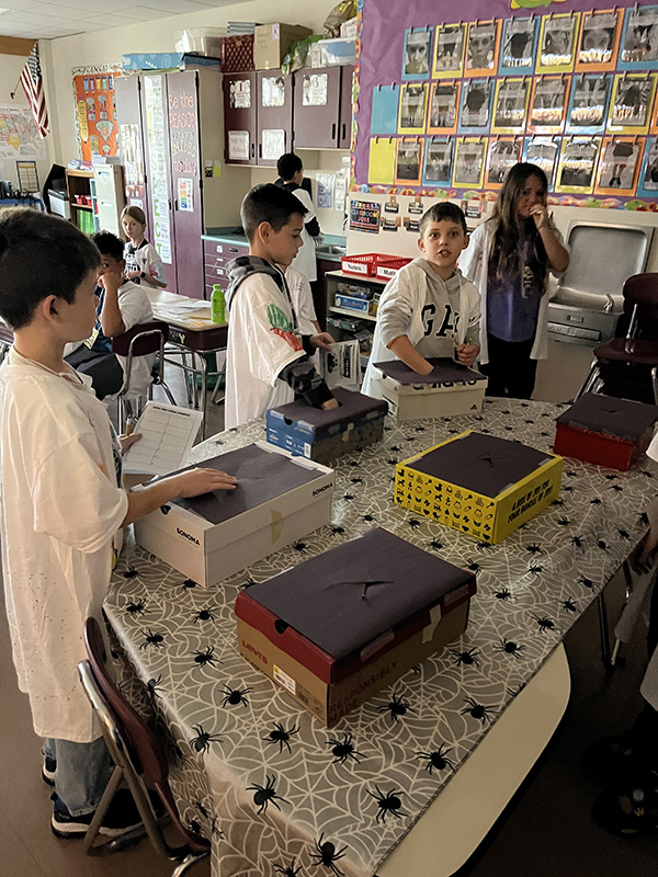 Several fourth-grade students stand around a table and put their hands into boxes.