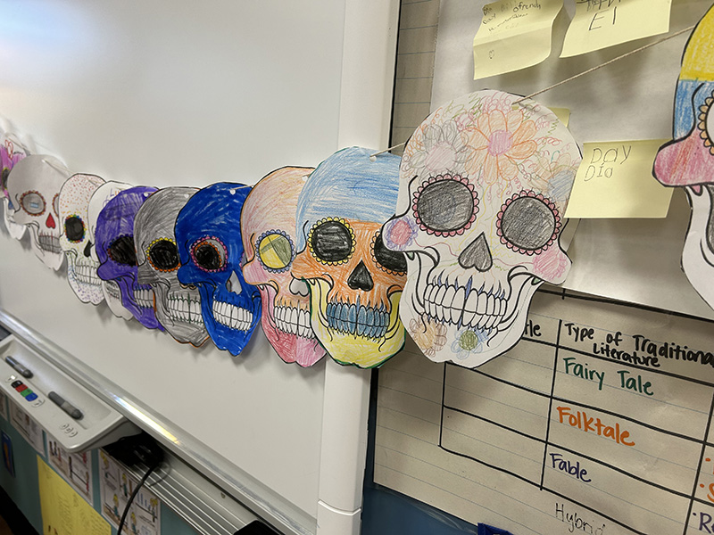 Paper skulls hang from a line on a wall. They are colored many different colors.