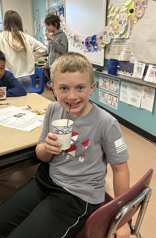 A boy with short blonde hair smiles as he sips from a cup.