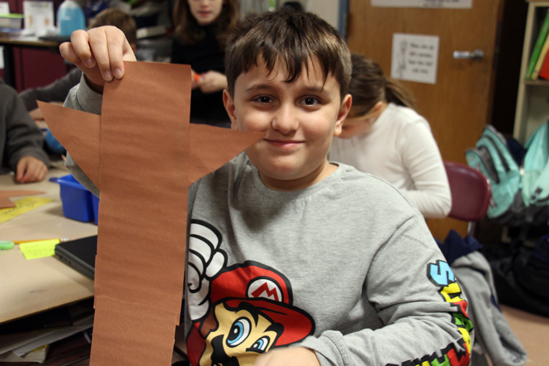 A fourth-grade boy with short brown hair smiles as he holds up a cutout brown totem pole.