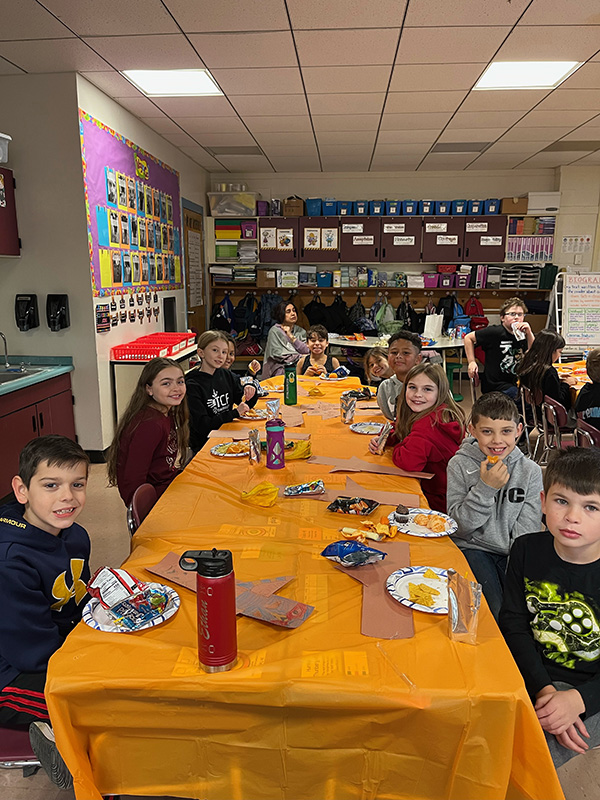 Long tables with orange tablecloths. Kids are sitting at them with snacks.