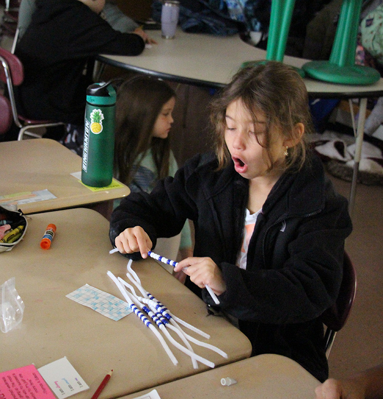 A fourth-grade girl sits at a desk with a happily surprised look on her face. She is putting blue and white beads on white plastic strings.