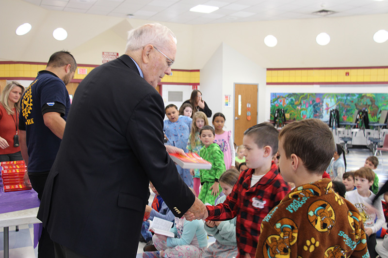 A man in a black suit jacket shakes the hand of a boy as he hands him a red dictionary.