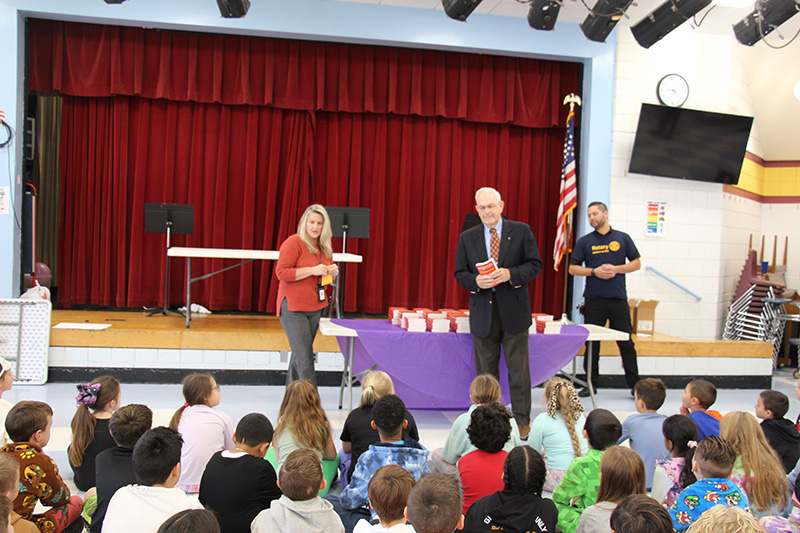 A woman stands to the left in front of a large group of elementary students. A man on the right, wearing a suit jacket, waits to talk.