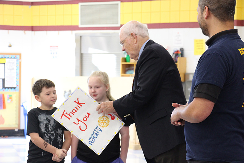 A man in a black suit jacket accepts a large card that says Thank you on it from two third-grade students.