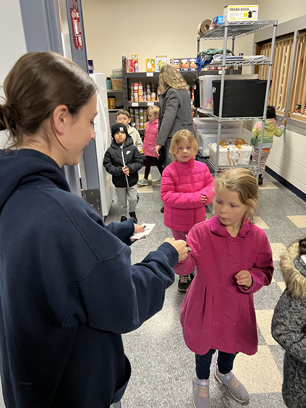 A line of kindergarten children walk past a young woman who is giving them each a sticker.