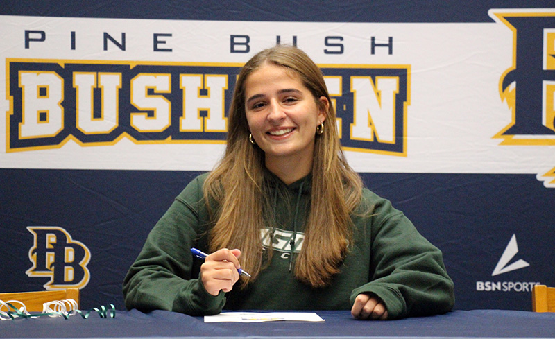 A young woman with long blonde hair smiles as she signs a piece of paper on the table in front of her. Behind her says Pine Bush Bushmen.