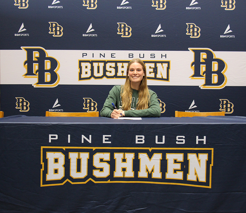 A young woman with long blonde hair sits at a table signing a piece of paper. She is wearing a green shirt. The tablecloth says Pin eBush Bushmen.