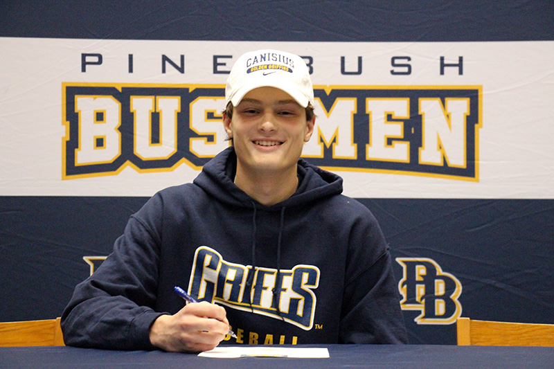 A young man smiles broadly as he signs a piece of paper on a desk. He is wearing a white baseball cap and a sweatshirt that says Griffs.