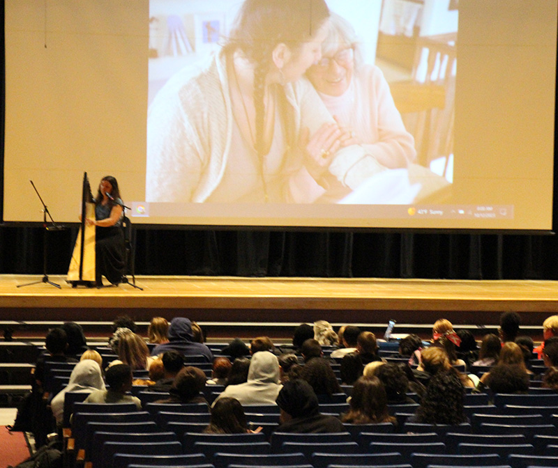 An auditorium of high school students sitting in chairs. There is a woman on the stage playing the harp and a screen with a young women and an older woman on it.