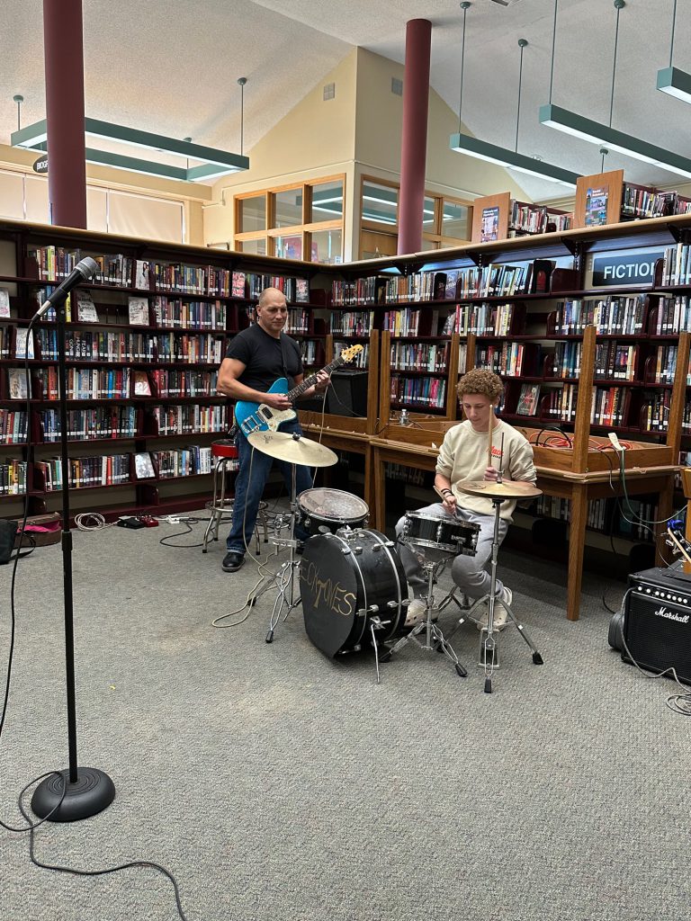 Two men play a guitar and drums in the library.