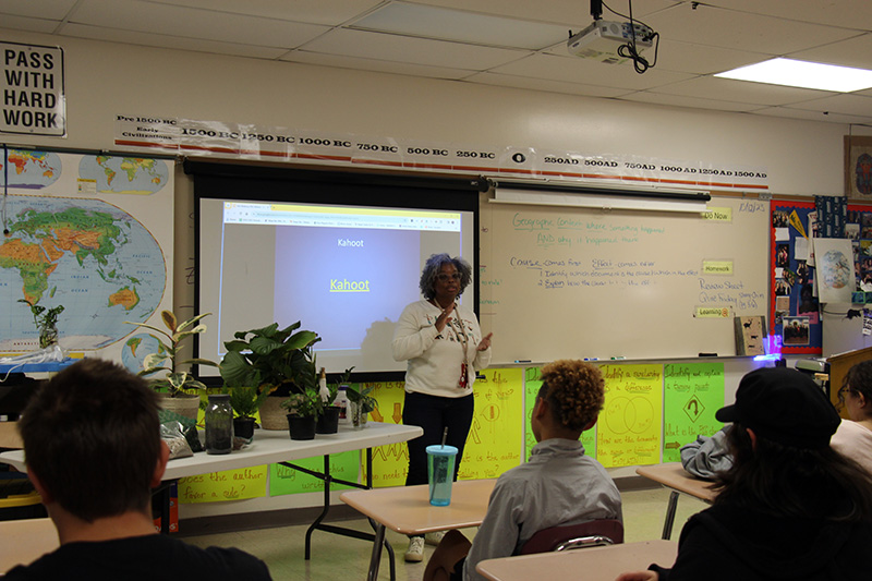 A woman stands at the front of a classroom with plants.