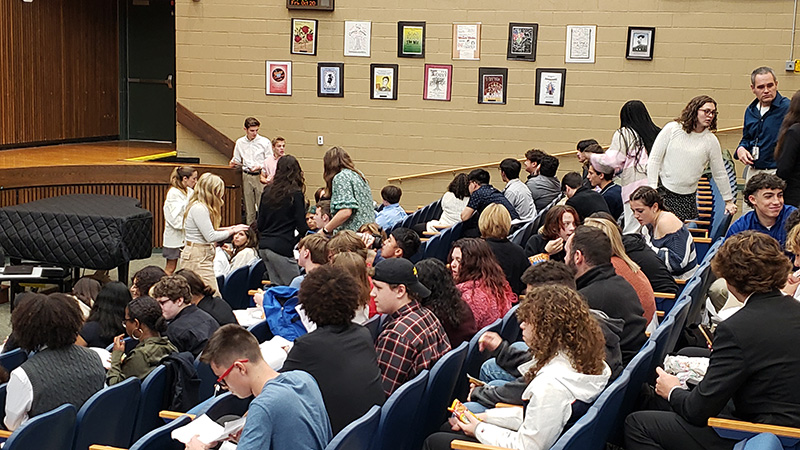 Taken from the back of an auditorium, there are many kids sitting in seats and some standing in front.