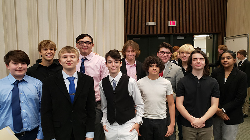 Eleven high school students stand in two rows. 10 boys and one girl. They are dressed very nicely, in jackets and ties.