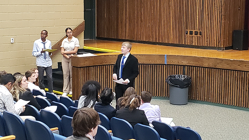 In an auditorium, kids are sitting in seats and a young man is in front presenting.