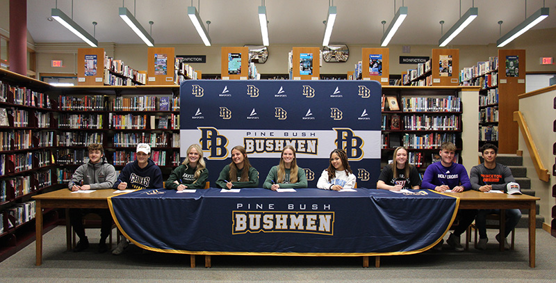 A group of nine high school students sit at a long table with a tablecloth that says Pine Bush Bushmen. Behind them is a background of blue and gold and white that has PB and Bushmen on it.