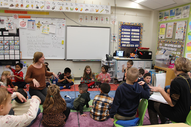 A group of kindergarten students sit in a big circle on the floow with two adults.