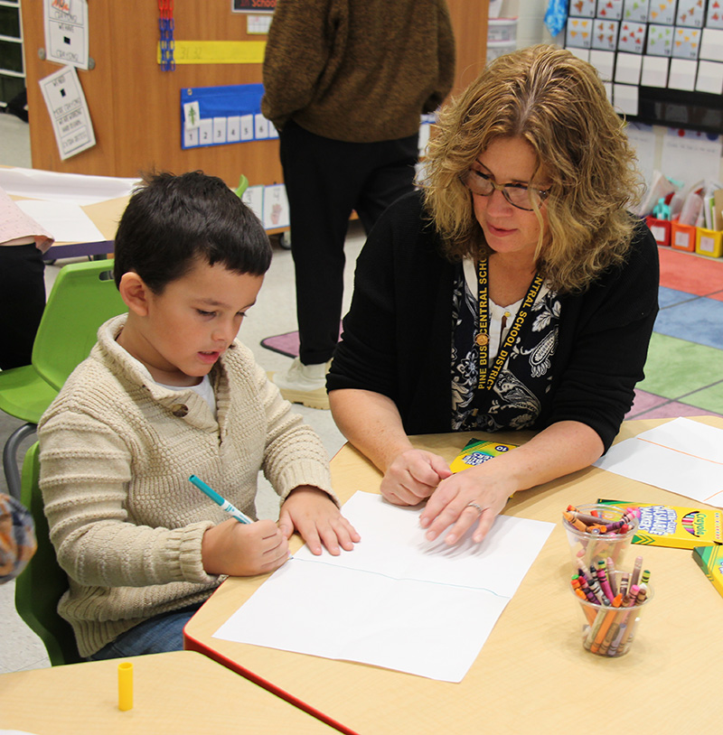 A woman with long light hair and eyeglasses sits at a desk with a kindergarten student as he works on his project. He has short dark hair and a is wearing a tan sweater.