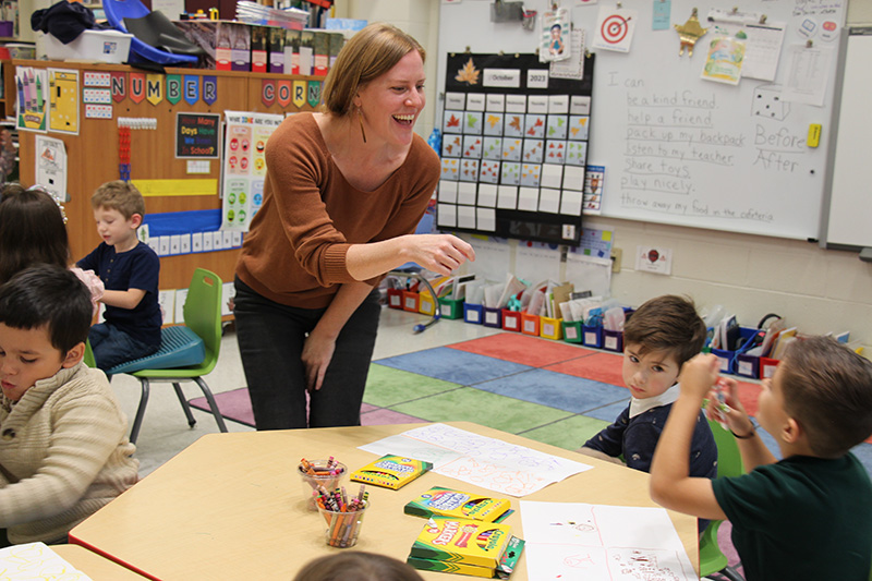 A woman has a big smile as she talks to kindergarten kids sitting at a table.