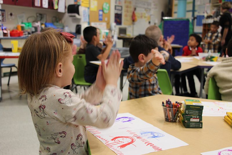 A group of kindergarten kids sitting at a table clap happily.