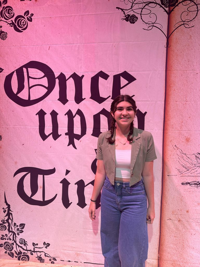 There is a pink background with a big book that says once upon a Time. In front is a high school girl smiling. She has shoulder-length  braids and is wearing jeans, a white shirt and tan overshirt.