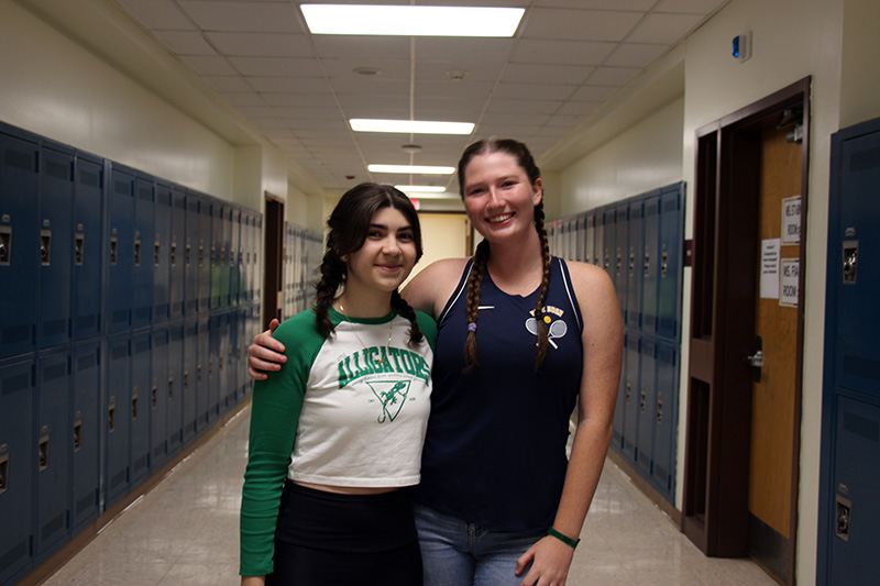 Two high school girls stand together smiling. They both have braids. Behind them is a school hallway lined with lockers.