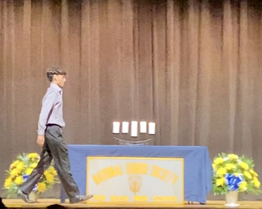 A young man walks across a stage by a table with five candles on it. The tablecloth says National Honor Society Pine Bush High School.