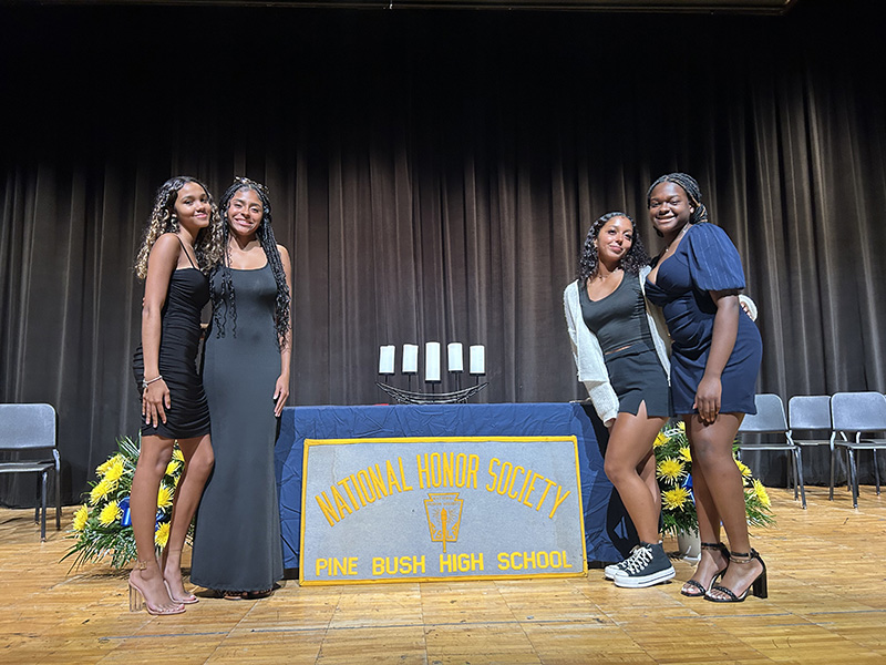 Four high school girls dressed in black dresses stand around a table with a cloth that says National Honor Society Pine Bush High School. On the table are five candles.