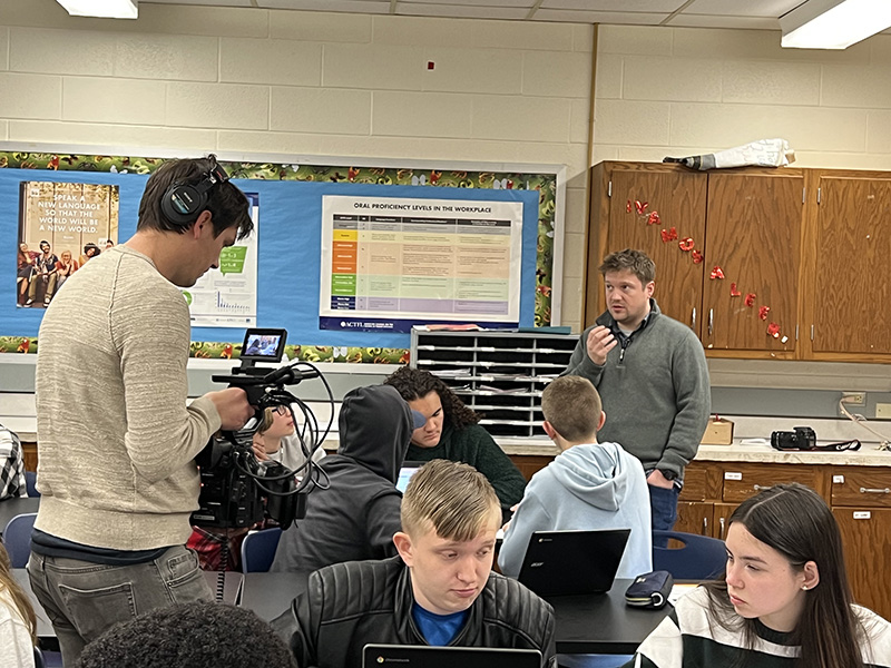 A man holds a camera as he videos a man teaching a group of high school students.