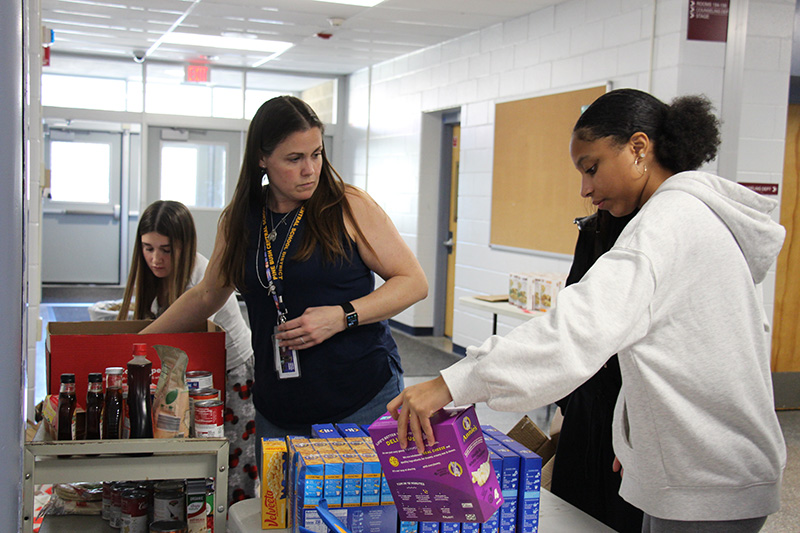 A woman in a blue shirt looks at a table filled with boxed food. A high school student is sorting the food.