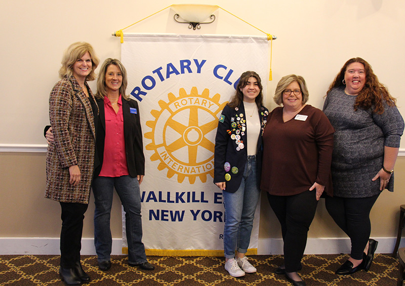 Four women and a high school girl stand together smiling. In the background is a banner that says Rotary Club Wallkill New York.